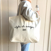 Alpha Chi Omega – A stylish college student wears an Alpha Chi Omega tote bag over her shoulder, proudly displaying the sorority’s name and founding year.