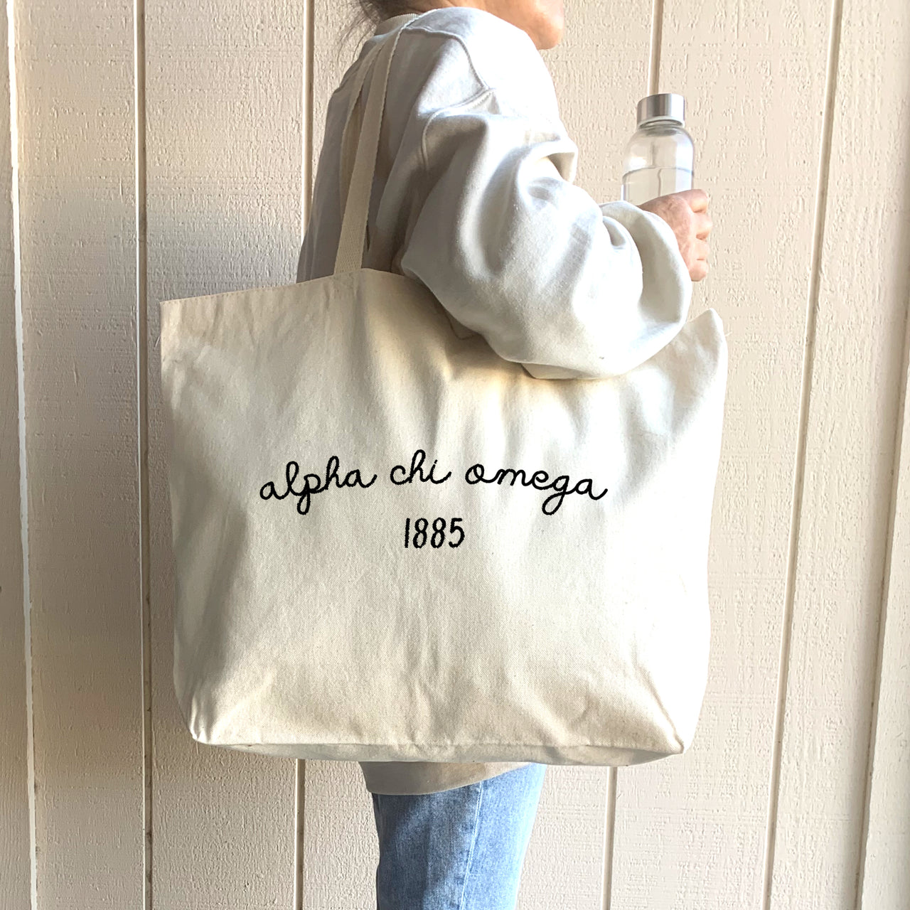 A college student confidently carries her Alpha Chi Omega tote over her shoulder, featuring the sorority’s name and founding year.