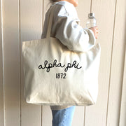 Alpha Phi – A confident college student carries her Alpha Phi tote bag across campus, a stylish and practical accessory for Greek life.