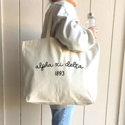 Alpha Xi Delta – A sorority sister beams while wearing an Alpha Xi Delta tote bag, a must-have for Greek life events and daily use.