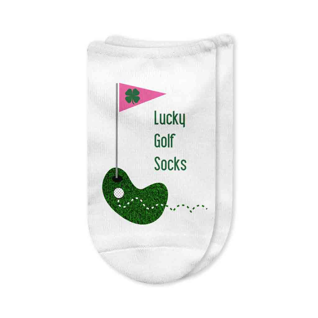 A pair of white no-show golf socks with the phrase "Lucky Golf Socks" printed on top, featuring a design of a golf ball rolling toward the hole with a pink flag and a four-leaf clover. A fun and stylish golf accessory for women.