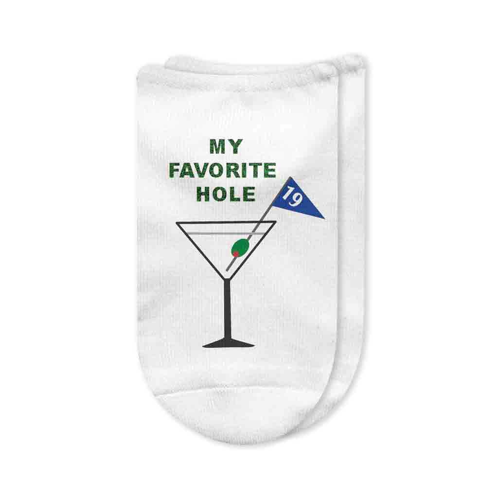 A pair of white no-show golf socks featuring the humorous phrase "MY FAVORITE HOLE" printed on top, along with a cocktail glass and a 19th hole flag piercing an olive. A fun and stylish gift for golfers who enjoy post-game drinks.